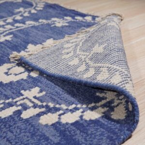 This blue rug with white patterns is folded over, revealing its woven backing and fringe near wood flooring.
