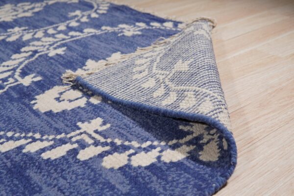 This blue rug with white patterns is folded over, revealing its woven backing and fringe near wood flooring.