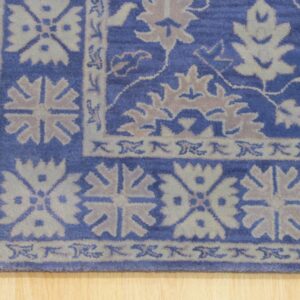 Corner view of a blue rug with complex beige and gray border pattern resting on a light wood floor.