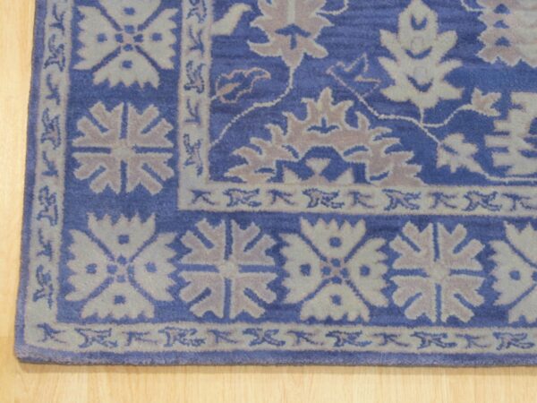 Corner view of a blue rug with complex beige and gray border pattern resting on a light wood floor.