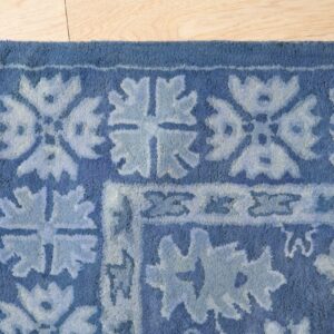 Corner view of a plush blue rug featuring a stylized light blue floral border pattern on light wood flooring.
