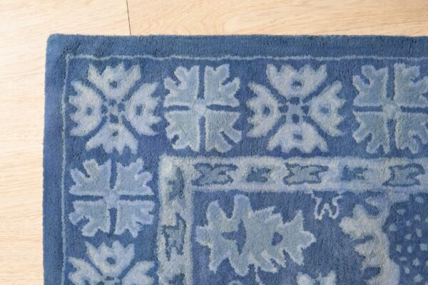 Corner view of a plush blue rug featuring a stylized light blue floral border pattern on light wood flooring.