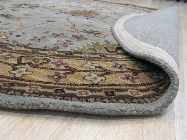 Traditional light blue and gold floral rug partially rolled back, showing its woven backing on light wood flooring.