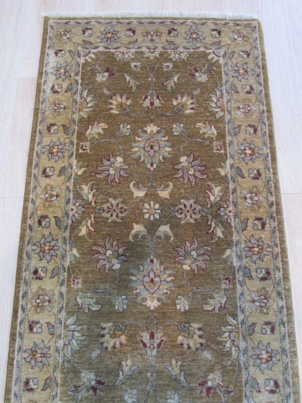 Muted olive runner rug with a repeating floral pattern and a beige border, displayed on pale wood planks.