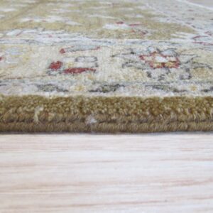 The thick edge of a cream and olive patterned rug with red accents rests on light wood flooring.