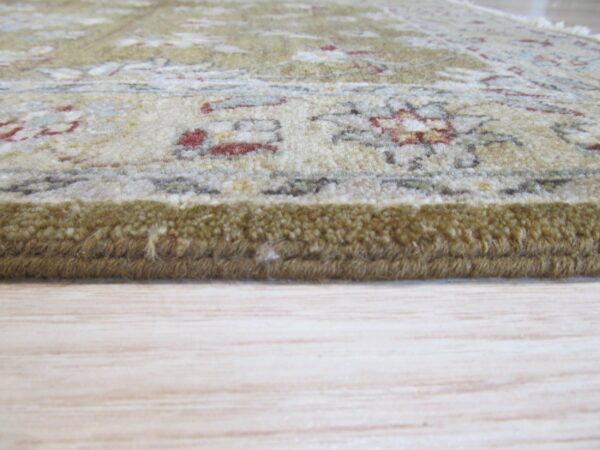 The thick edge of a cream and olive patterned rug with red accents rests on light wood flooring.