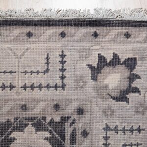 Close-up of a low-pile rug with geometric and floral patterns in beige, gray, and dark charcoal, featuring short fringes on light wood flooring.