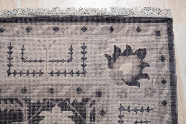 Close-up of a low-pile rug with geometric and floral patterns in beige, gray, and dark charcoal, featuring short fringes on light wood flooring.
