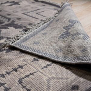 A gray and taupe geometric rug with fringe is partially rolled back on light wood flooring.