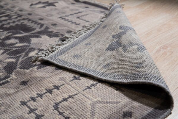 A gray and taupe geometric rug with fringe is partially rolled back on light wood flooring.