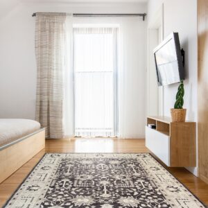 A charcoal and beige traditional patterned rug rests on hardwood floors in a sunlit bedroom.