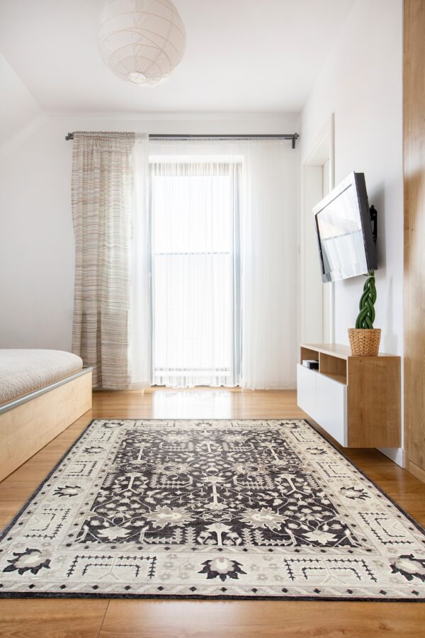 A charcoal and beige traditional patterned rug rests on hardwood floors in a sunlit bedroom.