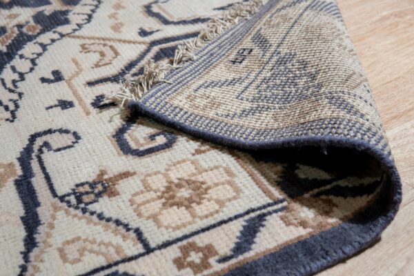 Close-up of a cream and charcoal patterned rug corner lifted on wood floor, showing its fringe, woven backing, and pile height.