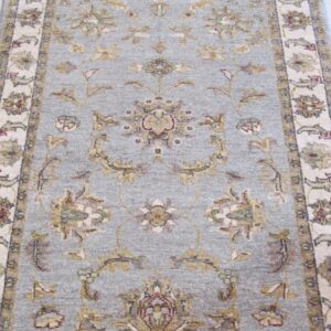 Muted blue-gray rug with an ornate traditional floral pattern and pale fringed border on light wood flooring.