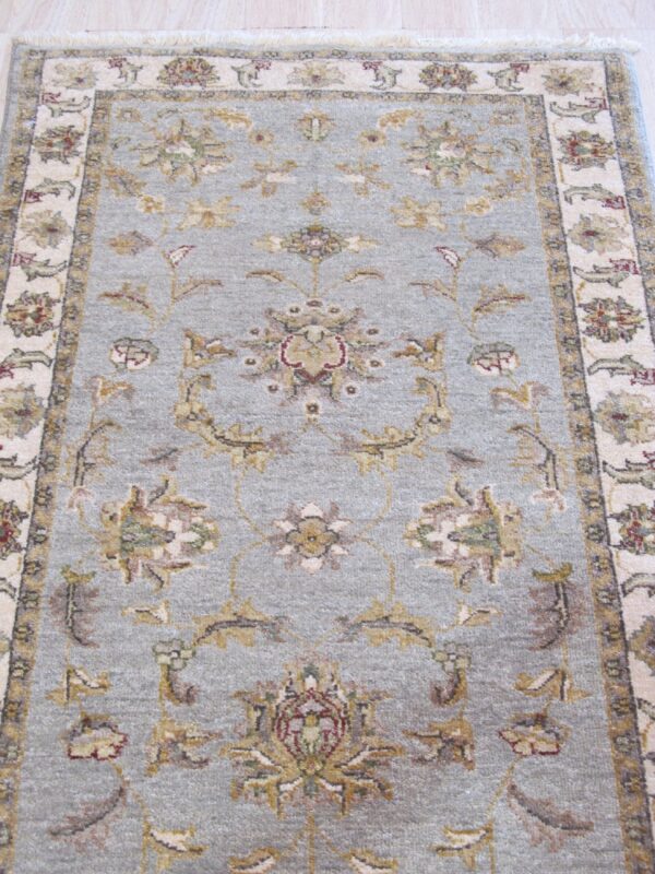 Muted blue-gray rug with an ornate traditional floral pattern and pale fringed border on light wood flooring.