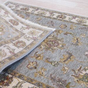 Close-up of a blue-gray traditional rug with fringe, partially rolled back on light wood planks.
