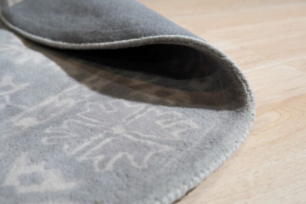 The curled edge of a low-pile gray rug with a light patterned design rests on blonde wood flooring.