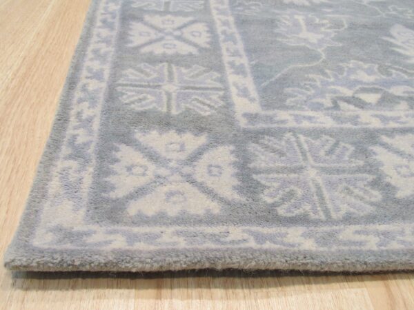 Angled view of a plush gray rug with white traditional border design resting on light wood plank flooring.