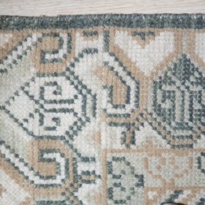 Textured rug corner displaying geometric patterns in cream, tan, and dark green on light flooring.