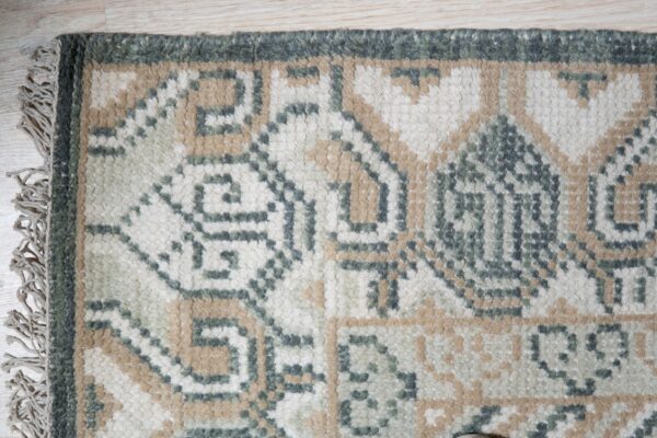Textured rug corner displaying geometric patterns in cream, tan, and dark green on light flooring.