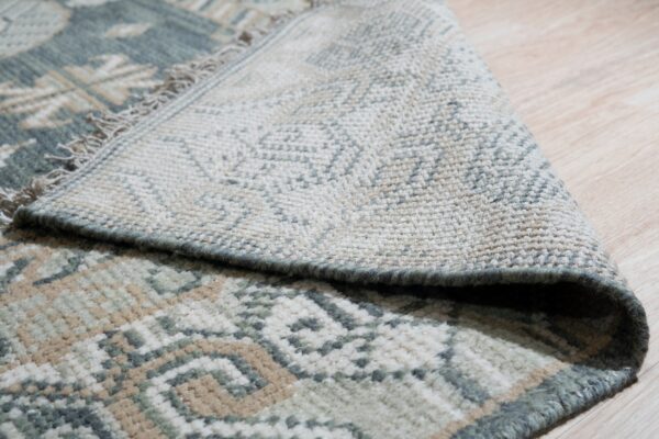 Detail view of a low-pile rug in sage, tan, and cream, showing the fringe and woven reverse side.