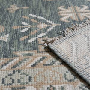 The low pile, green and beige geometric rug is folded back revealing the woven backing and tassels.
