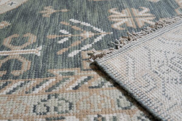 The low pile, green and beige geometric rug is folded back revealing the woven backing and tassels.