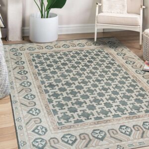 Teal and cream ornate geometric rug on wood floors beside a large plant and white armchair.