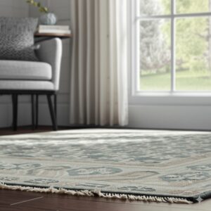 A low-pile teal and tan traditional rug with fringe covers dark wood flooring in a bright room.