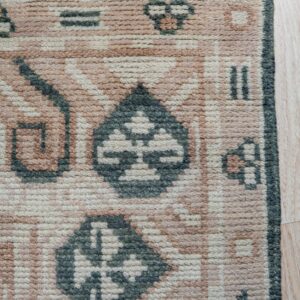 Corner detail of a textured pink, cream, and green geometric rug with fringe on light wood.