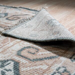 A low-pile rug with tan and sage geometric patterns is folded back, revealing the backing on light wood flooring.