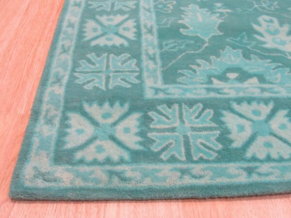 Textured teal and aqua rug displaying traditional borders and scrolling floral patterns on pinkish wood planks.