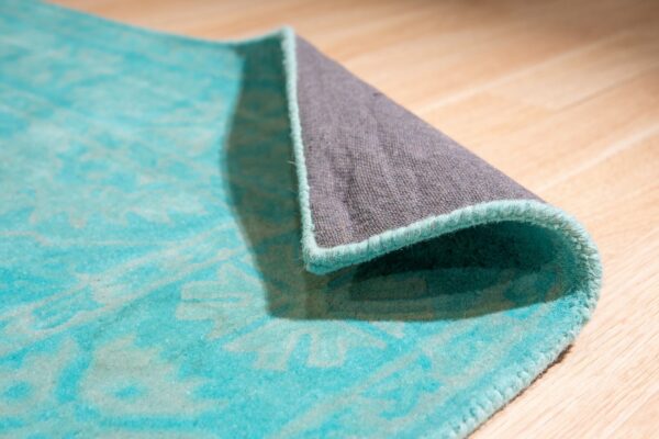 Turquoise low-pile rug with a pale pattern is rolled, showing the gray backing on light wood.