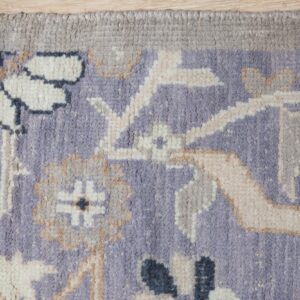 A medium-pile rug featuring an ornate lavender, cream, and gray floral pattern and fringed edge on wood flooring.