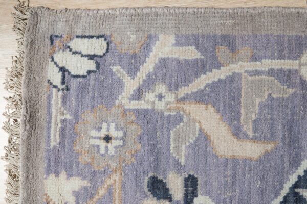 A medium-pile rug featuring an ornate lavender, cream, and gray floral pattern and fringed edge on wood flooring.
