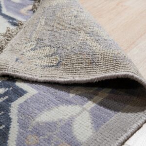 A cream, purple, and grey patterned rug is rolled back, revealing its woven underside on light wood flooring.