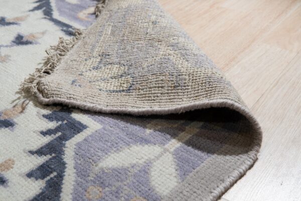 A cream, purple, and grey patterned rug is rolled back, revealing its woven underside on light wood flooring.
