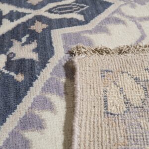 Detailed view of a blue, cream, and lavender patterned rug, folded back to reveal the woven reverse and fringe.