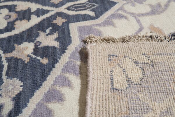 Detailed view of a blue, cream, and lavender patterned rug, folded back to reveal the woven reverse and fringe.
