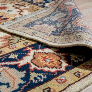 Cream, navy, and rust patterned rug detail showing the woven backing curled on a light wood floor.