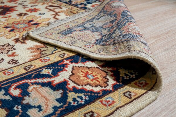 Cream, navy, and rust patterned rug detail showing the woven backing curled on a light wood floor.