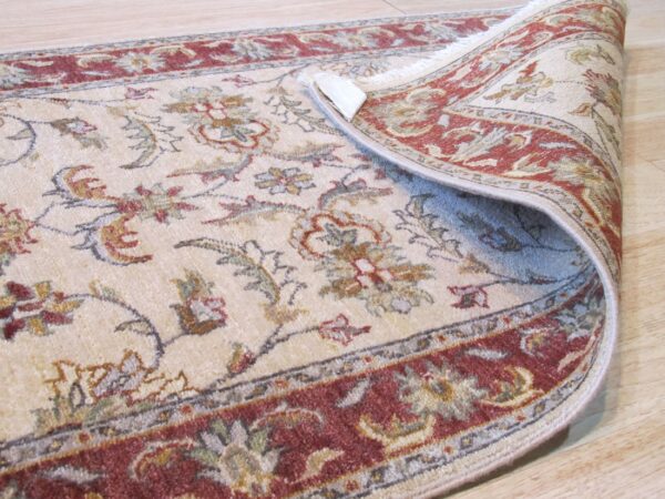 The corner of a light-colored rug with a red floral border is curled back on light wood flooring.