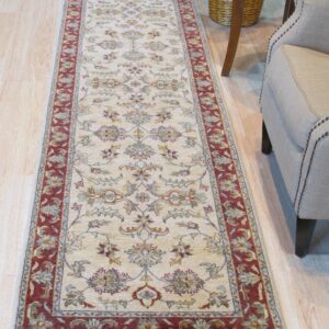 Cream floral runner rug with a red border lies on light wood floors beside a tan armchair.