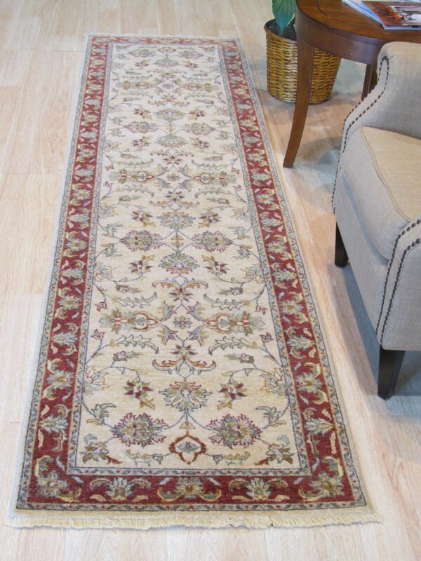 Cream floral runner rug with a red border lies on light wood floors beside a tan armchair.