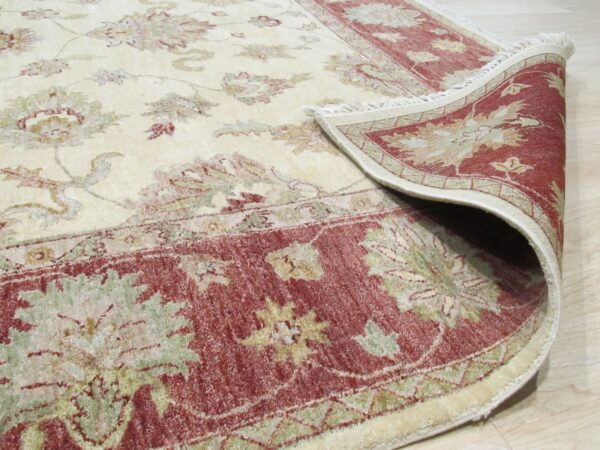 Beige floral rug with red and sage green botanical motifs, shown folded on light wood flooring.