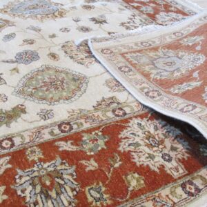A cream rug with rust border and muted floral patterns is partially rolled back on light wood flooring.