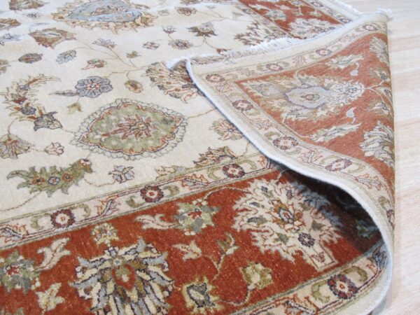 A cream rug with rust border and muted floral patterns is partially rolled back on light wood flooring.