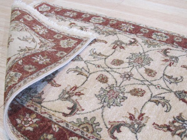 Cream low-pile rug with rust borders and muted green floral vine pattern rests on wood flooring.