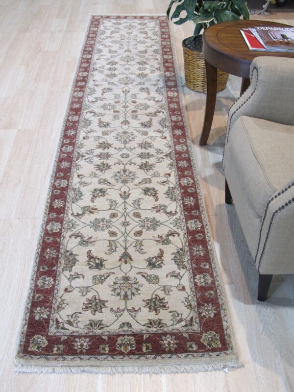 Cream runner rug with red border and detailed floral pattern on pale wood near an upholstered chair.