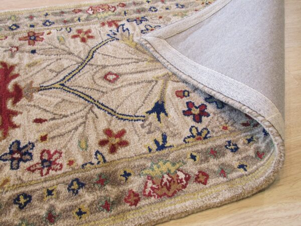 Textured tan rug with a traditional floral design, folded over to expose the gray woven backing on wood flooring.